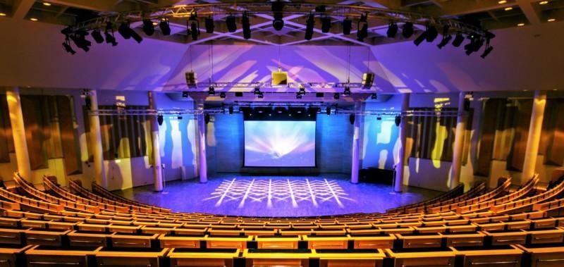 Picture of Lappeenranta Hall. Several long and empty rows of benches with an empty area in front of them for the performances. The hall is lit with different colored lights.