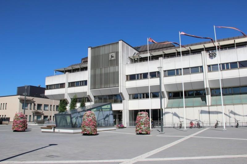 Picture of the town hall. It is a large and multi-storey building with a light facade. In front of the building is a large square, floral arrangements and several flagpoles. There is also another building next to the town hall.